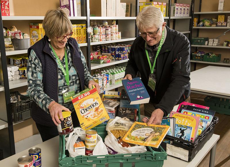 A man and woman adding books and food to a shopping bag with shelves of dry goods behind them